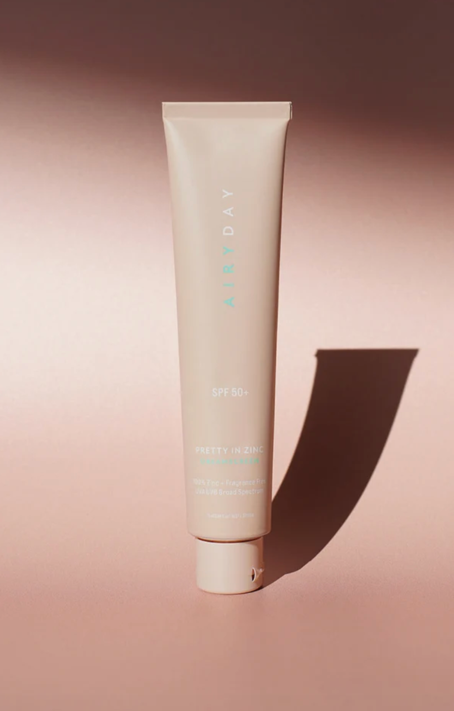 A tube of AIRDAY Pretty in Zinc SPF 50+ sunscreen, standing upright on a warm pinkish-beige background with a sharp shadow cast to the right.