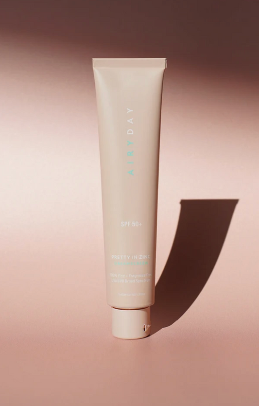 A tube of AIRDAY Pretty in Zinc SPF 50+ sunscreen, standing upright on a warm pinkish-beige background with a sharp shadow cast to the right.